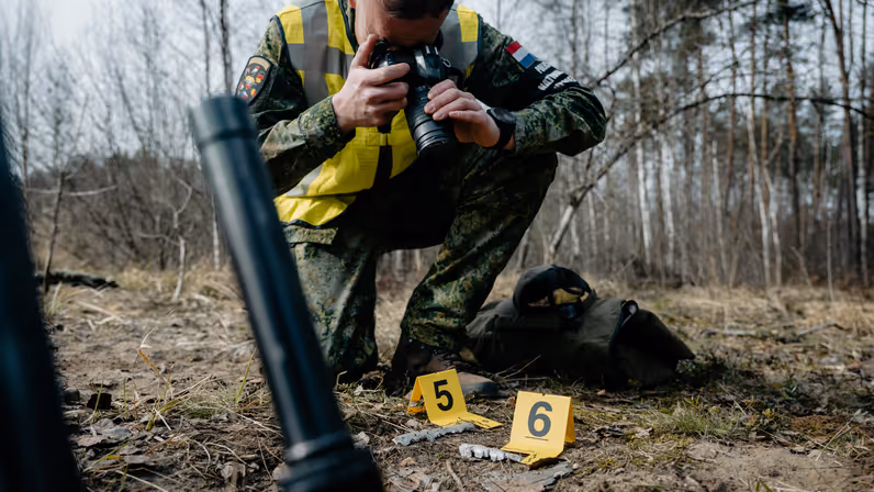 Een marechaussee die een bewijsstuk fotografeert. Op de grond staan een aantal bordjes met daarop een nummer.  