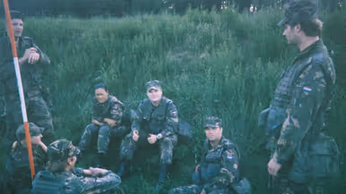 Een jonge Tim Nelissen in gevechtstenue zittend in het hoge gras samen met zes andere militairen, die op hem heen zitten en staan. Tim steekt beide duimen op richting de camera.