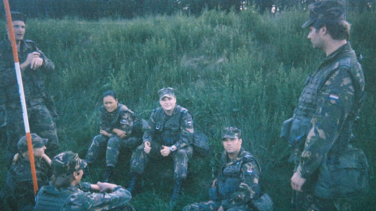 Een jonge Tim Nelissen in gevechtstenue zittend in het hoge gras samen met zes andere militairen, die op hem heen zitten en staan. Tim steekt beide duimen op richting de camera.