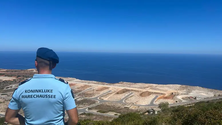 Een marechaussee, vanaf de rug gezien, kijkt vanaf een hoog punt uit over een oefenterrein en een stuk zee.