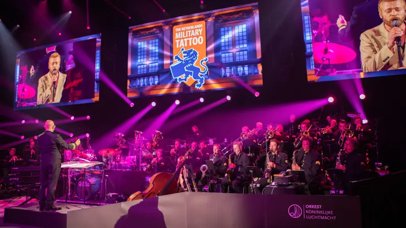 Het Orkest Koninklijke Luchtmacht speelt muziek onder paarse podiumverlichting. Erboven hangen drie televisieschermen met op de buitenste twee dezelfde zanger en in het midden de tekst ‘The Netherlands Military Tattoo’.