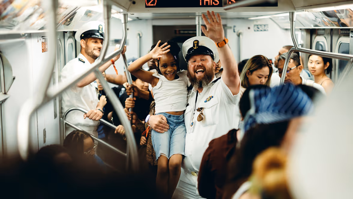  Een marineman in de metro, die een meisje op zijn arm heeft. Beiden zwaaien naar de camera. 