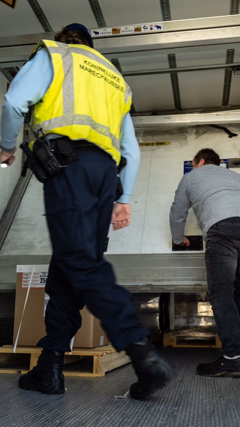 Een chauffeur schuift in een oplegger een paneel, weg dat voor de lading zit. Een marechaussee kijkt toe.