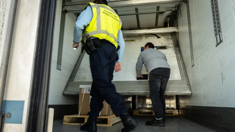 Een chauffeur schuift in een oplegger een paneel, weg dat voor de lading zit. Een marechaussee kijkt toe.