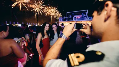 een marineman fotografeert twee mooi aangeklede dames, met op de achtergrond vuurwerk.&nbsp;