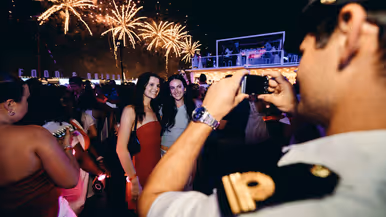 een marineman fotografeert twee mooi aangeklede dames, met op de achtergrond vuurwerk.&nbsp;