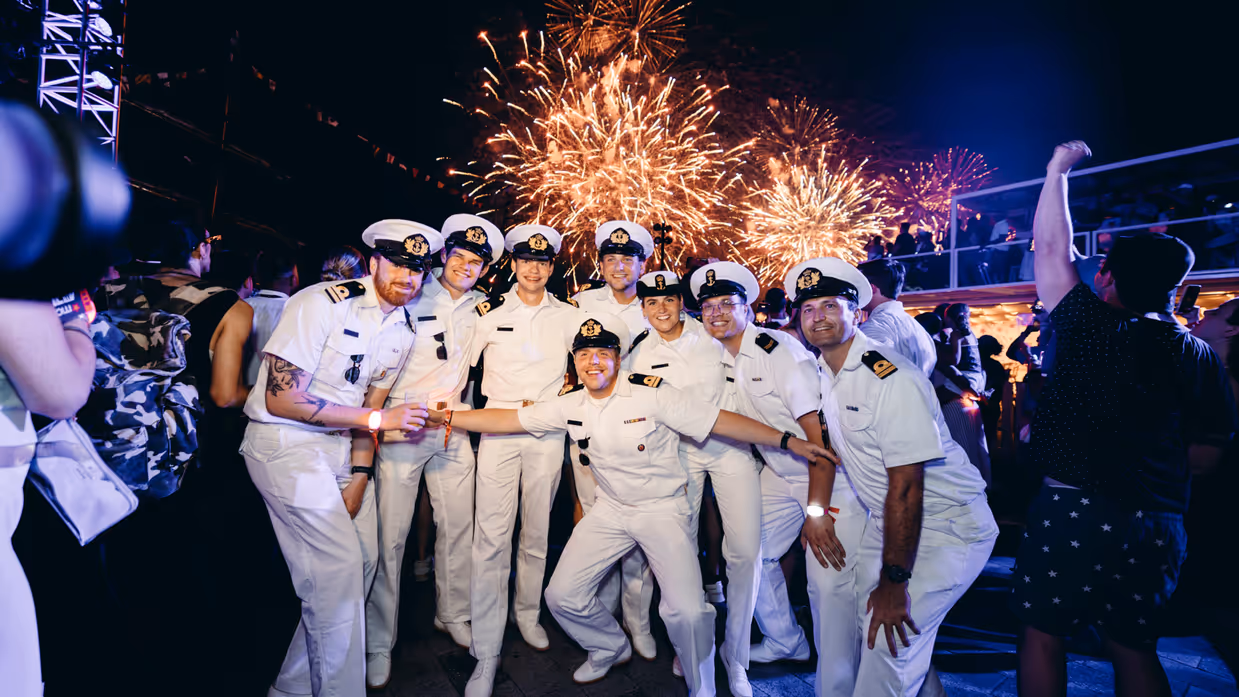 een poserend groepje opvarenden in wit uniform met op de achtergrond vuurwerk.  