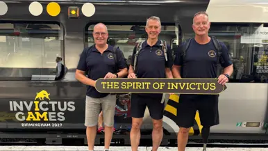 Drie mannen in korte broek, sportschoenen en een donkerblauw shirt houden een bord vast met de tekst ‘The spirit of invictus.’ Achter hen staat een metro met daarop de tekst ‘Invictus Games Birmingham 2027’.