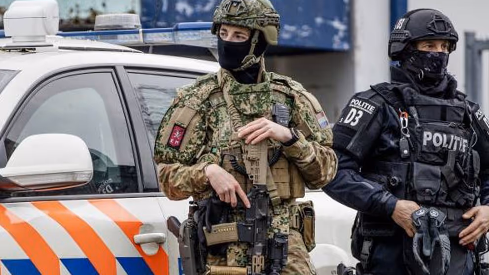 En gewapende, gemaskerde militair staat voor een politieauto en naast een politieman met gezichtsbedekking.
