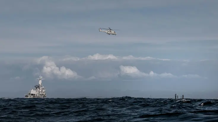 Het M-fregat Zr.Ms. Van Amstel op zee, een NH90 daarnaast in de lucht en een deel van de onderzeeboot net boven water.