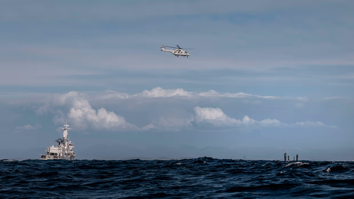 Het M-fregat Zr.Ms. Van Amstel op zee, een NH90 daarnaast in de lucht en een deel van de onderzeeboot net boven water.