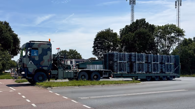 een groene trekker-oplegger beladen met veldbedden op een kruising.  
