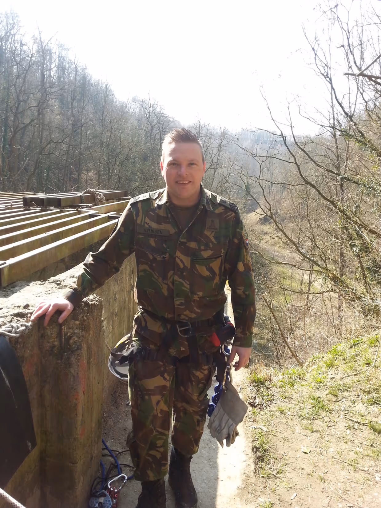 Een jonge Tim Nelissen in gevechtstenue poseert voor de foto in het buitenterrein. Om zijn heupen draagt hij een veiligheidstuigje met daaraan een helm en handschoenen vastgeklikt. Op de grond ligt een touw met karabijnhaak.
