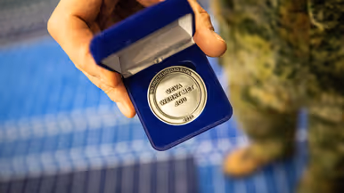 Een zilverkleurige munt met de tekst ‘Instructeursdag DCPL 2026; VeVa werkt met jou’. Deze wordt door een hand van een militair getoond in een blauw doosje.