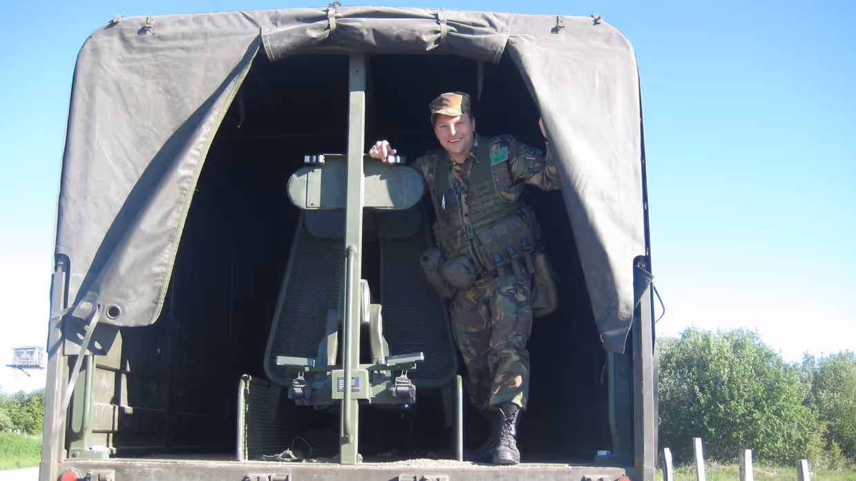 Een jonge Tim Nelissen in gevechtstenue poseert staand in de open achterkant van een militaire vrachtwagen en kijkt in de camera.