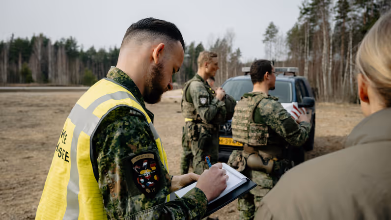 Een marechaussee op de rug gezien, die te velde en aantekening maakt. 