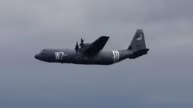 een C130 Hercules voorzien van geallieerde herkenningsstrepen tegen een donkere achtergrond.
