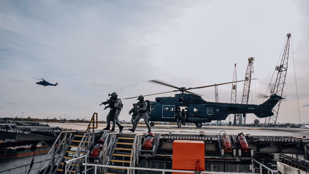 Apache-gevechtshelikopter op platform. Militairen met getrokken wapens ervoor. Verder weg een Apache in de lucht.
