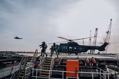 Apache-gevechtshelikopter op platform. Militairen met getrokken wapens ervoor. Verder weg een Apache in de lucht.