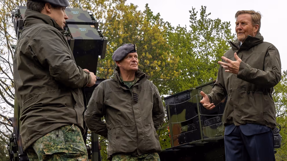 Koning Willem-Alexander in gesprek met 2 militairen. Ze staan voor een Patriot-luchtverdedigingssysteem.