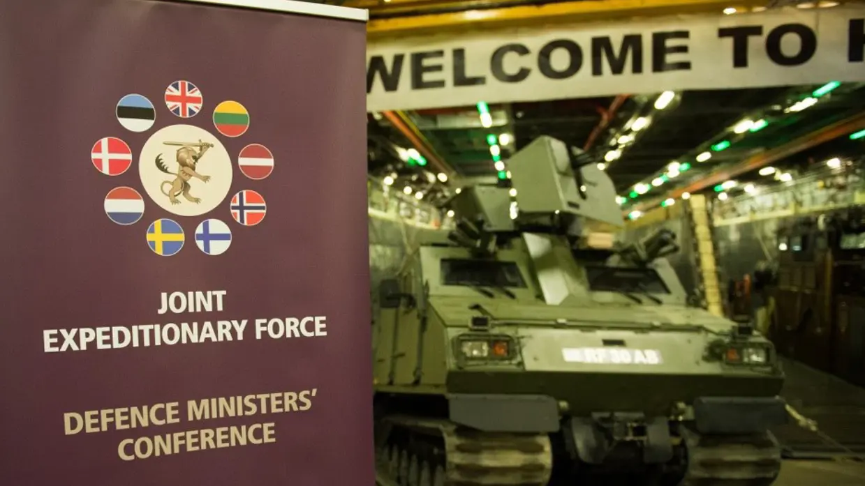 Een banner met daarop de tekst ‘Joint Expeditionary Force’ en de vlaggen van de deelnemende landen. Daarachter onscherp een militair voertuig.