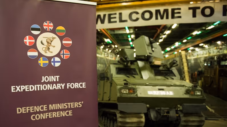 Een banner met daarop de tekst ‘Joint Expeditionary Force’ en de vlaggen van de deelnemende landen. Daarachter onscherp een militair voertuig.