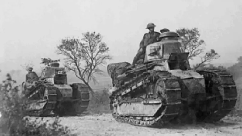 Twee oude tanks in een open veld met soldaten erop, omringd door bomen en struiken. 