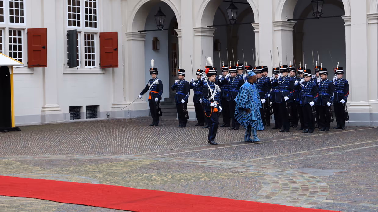links: Een vrouw in een prachtige, versierde mantel loopt over een rode loper, terwijl een groep geüniformeerde wachten in blauwe tenues met sabel en rode pluim haar salueren. Midden: Een man in een blauwe, traditioneel gewaad loopt langs een rij wachten in blauwe uniformen met sabels en pluimen, voor een wit gebouw met rode luiken en een rode loper. Rechts: Een man in een donkere jas loopt langs een rij wachten in blauwe uniformen met pluimen en sabels, voor een wit gebouw met rode luiken en ramen.