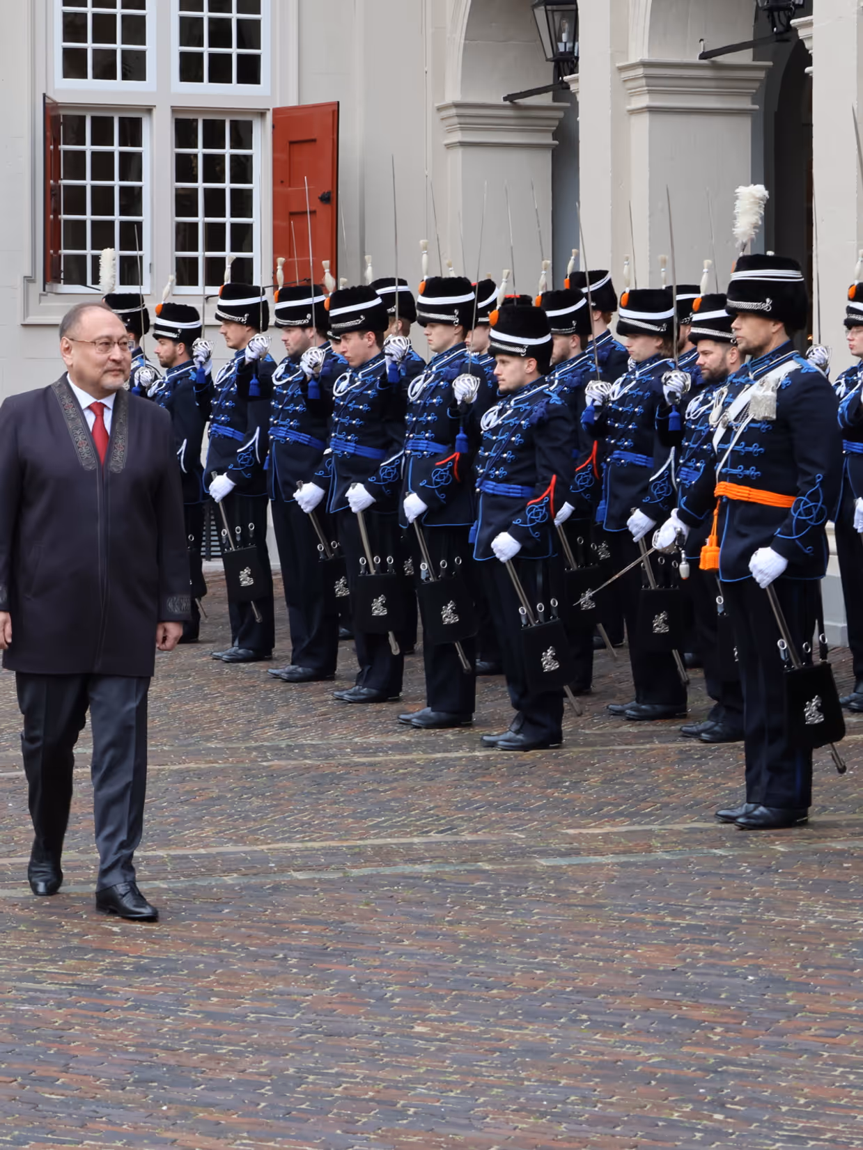 links: Een vrouw in een prachtige, versierde mantel loopt over een rode loper, terwijl een groep geüniformeerde wachten in blauwe tenues met sabel en rode pluim haar salueren. Midden: Een man in een blauwe, traditioneel gewaad loopt langs een rij wachten in blauwe uniformen met sabels en pluimen, voor een wit gebouw met rode luiken en een rode loper. Rechts: Een man in een donkere jas loopt langs een rij wachten in blauwe uniformen met pluimen en sabels, voor een wit gebouw met rode luiken en ramen.