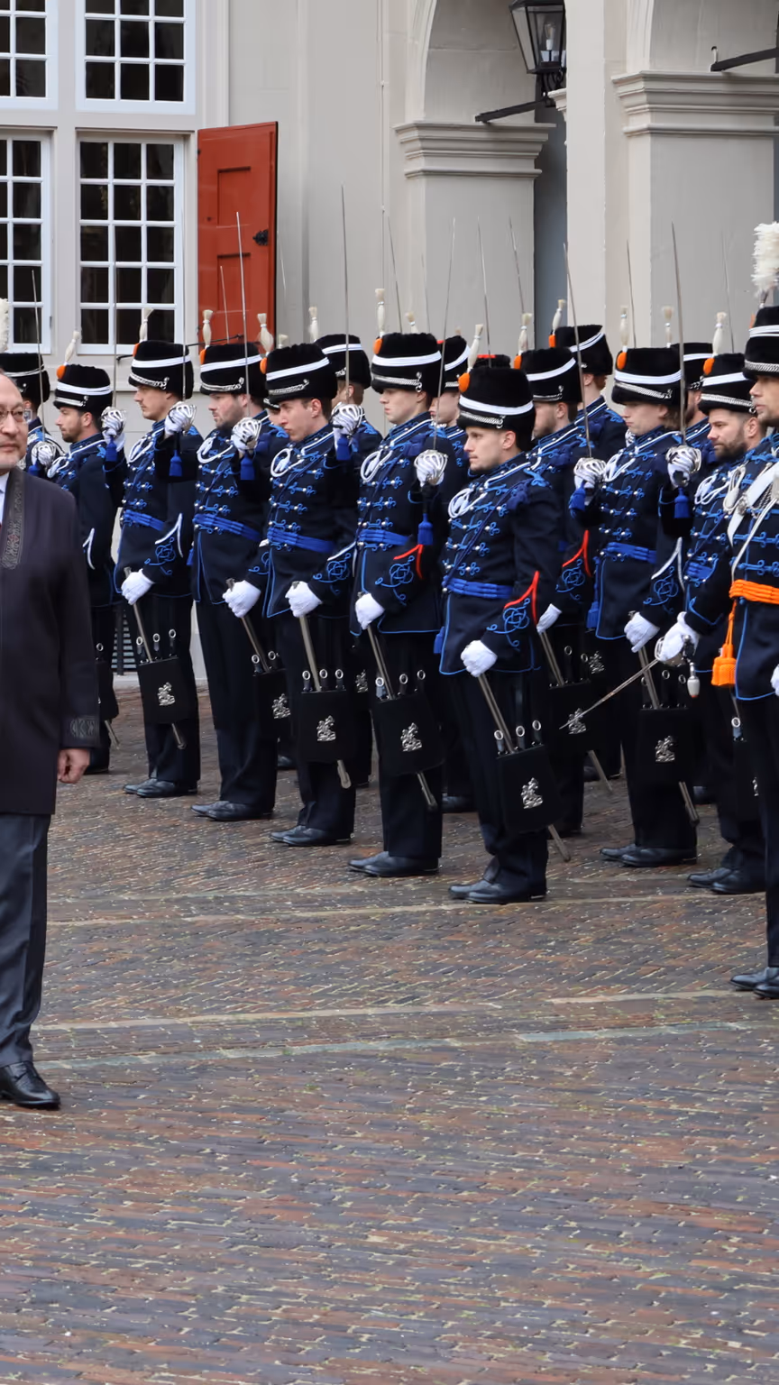 links: Een vrouw in een prachtige, versierde mantel loopt over een rode loper, terwijl een groep geüniformeerde wachten in blauwe tenues met sabel en rode pluim haar salueren. Midden: Een man in een blauwe, traditioneel gewaad loopt langs een rij wachten in blauwe uniformen met sabels en pluimen, voor een wit gebouw met rode luiken en een rode loper. Rechts: Een man in een donkere jas loopt langs een rij wachten in blauwe uniformen met pluimen en sabels, voor een wit gebouw met rode luiken en ramen.