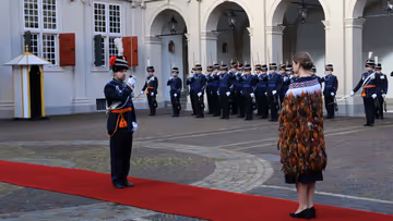 links: Een vrouw in een prachtige, versierde mantel loopt over een rode loper, terwijl een groep geüniformeerde wachten in blauwe tenues met sabel en rode pluim haar salueren. Midden: Een man in een blauwe, traditioneel gewaad loopt langs een rij wachten in blauwe uniformen met sabels en pluimen, voor een wit gebouw met rode luiken en een rode loper. Rechts: Een man in een donkere jas loopt langs een rij wachten in blauwe uniformen met pluimen en sabels, voor een wit gebouw met rode luiken en ramen.