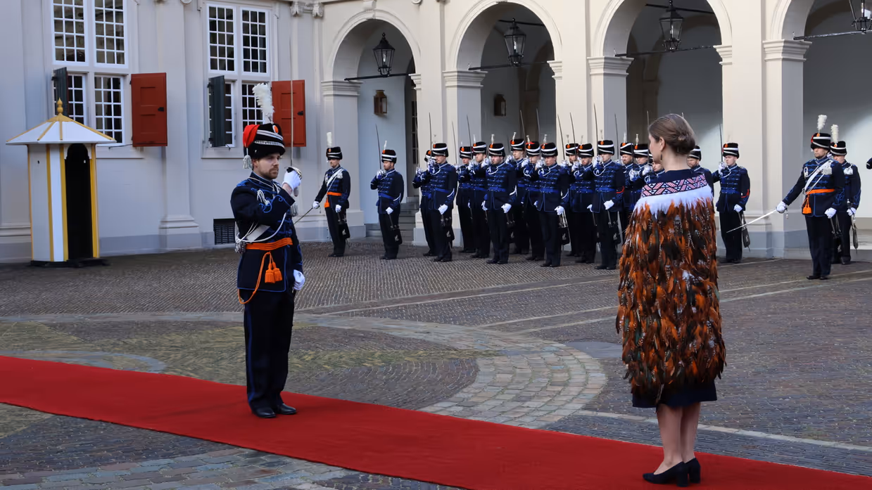 links: Een vrouw in een prachtige, versierde mantel loopt over een rode loper, terwijl een groep geüniformeerde wachten in blauwe tenues met sabel en rode pluim haar salueren. Midden: Een man in een blauwe, traditioneel gewaad loopt langs een rij wachten in blauwe uniformen met sabels en pluimen, voor een wit gebouw met rode luiken en een rode loper. Rechts: Een man in een donkere jas loopt langs een rij wachten in blauwe uniformen met pluimen en sabels, voor een wit gebouw met rode luiken en ramen.