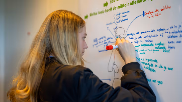 Een jonge vrouw schrijft met een rode marker op een wit vel papier. Daarop zijn meerdere teksten geschreven rondom een silhouet van een militair.