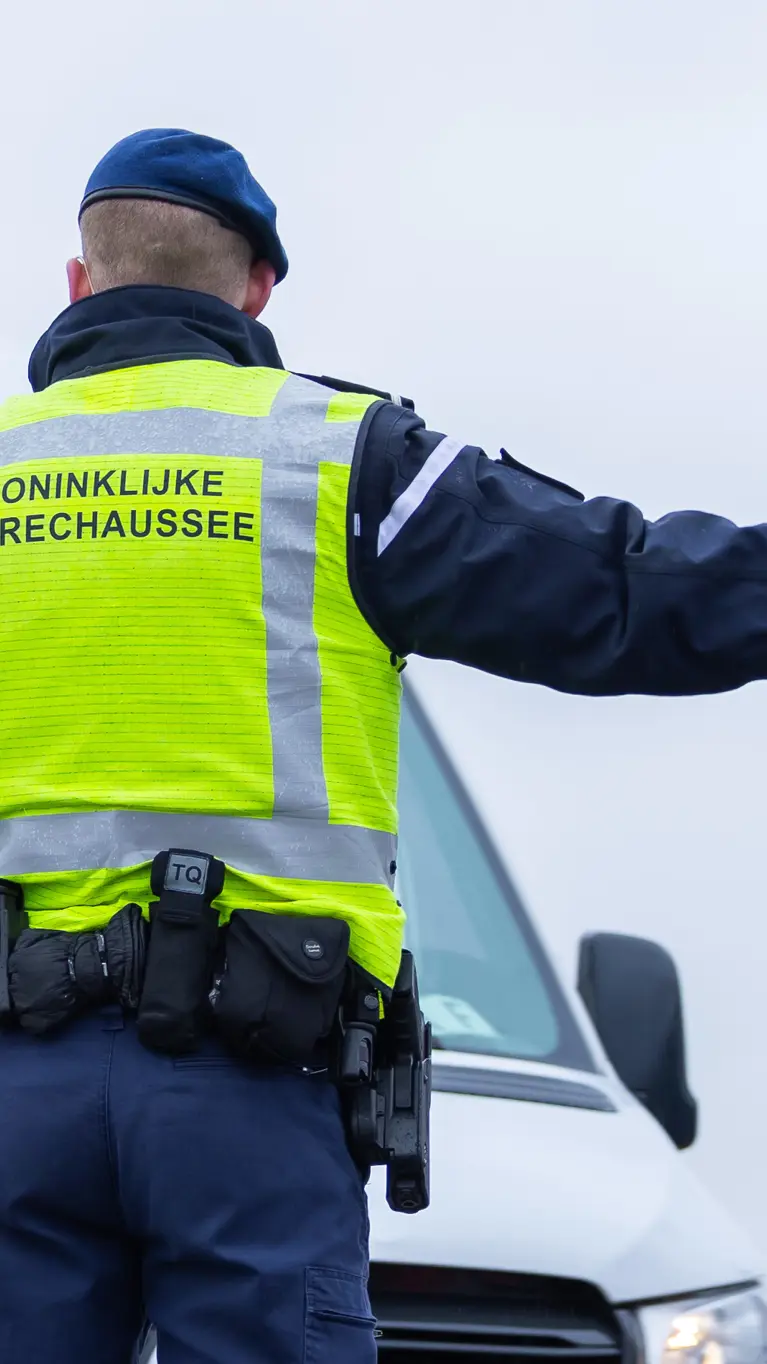 Foto van een marechaussee met een geel hesje. Hij wijst een auto de weg.