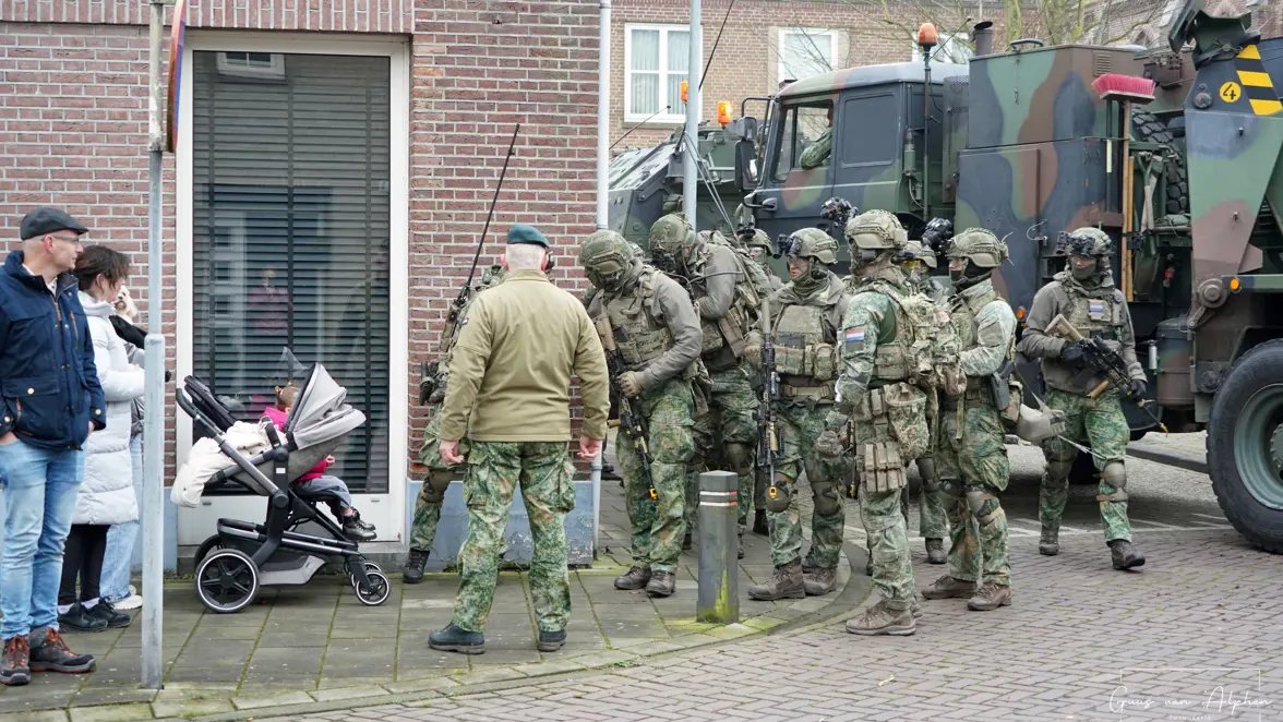 Zeven militairen met wapens staan naast een vrachtwagen en benaderen twee burgers in een gewone Nederlandse straat.