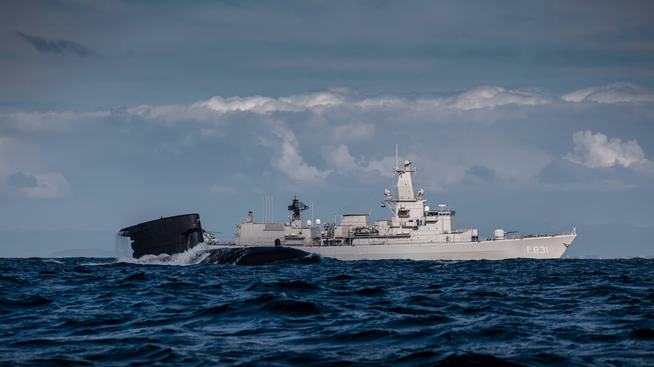 Het sail van een onderzeeboot dat half uit het water steekt met op de achtergrond het M-fregat Zr.Ms. Van Amstel.