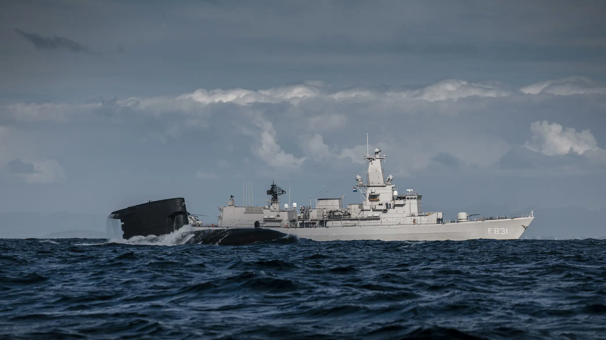 Het sail van een onderzeeboot dat half uit het water steekt met op de achtergrond het M-fregat Zr.Ms. Van Amstel.