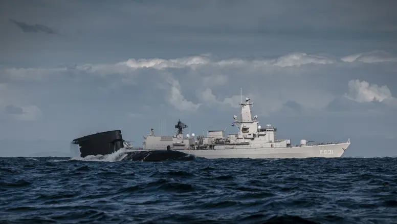 Het sail van een onderzeeboot dat half uit het water steekt met op de achtergrond het M-fregat Zr.Ms. Van Amstel.