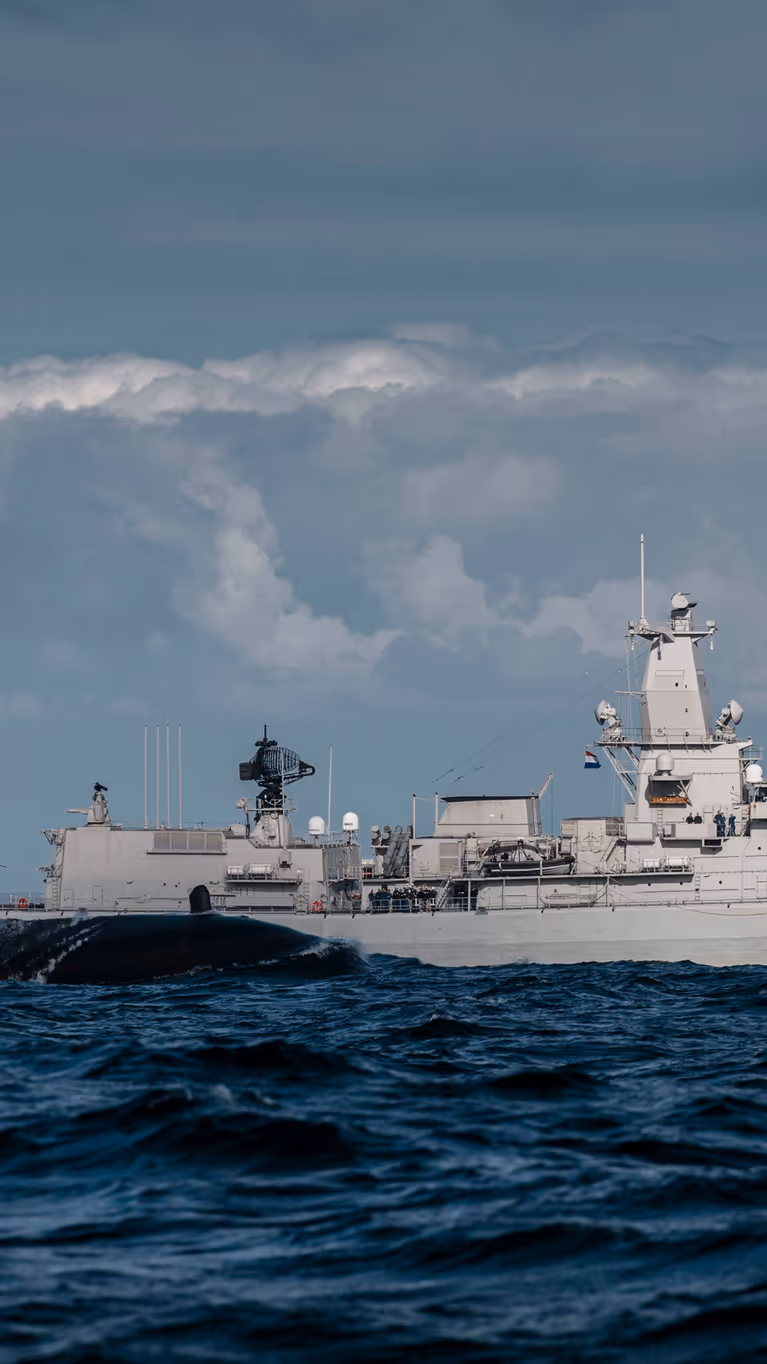 Het sail van een onderzeeboot dat half uit het water steekt met op de achtergrond het M-fregat Zr.Ms. Van Amstel.