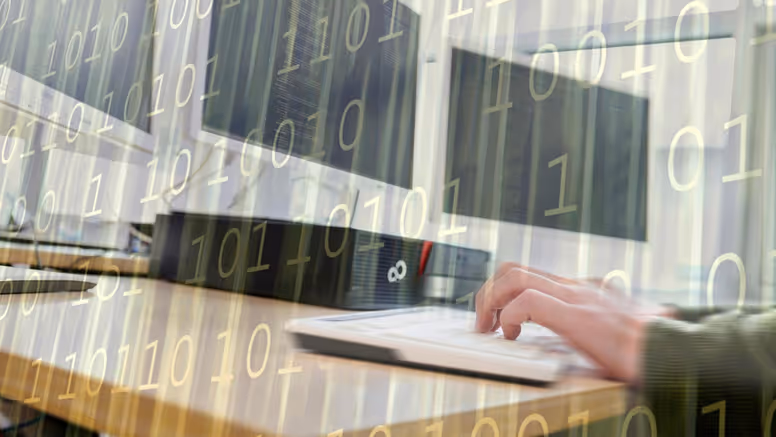 Een paar handen op een toetsenbord, achter computerschermen.