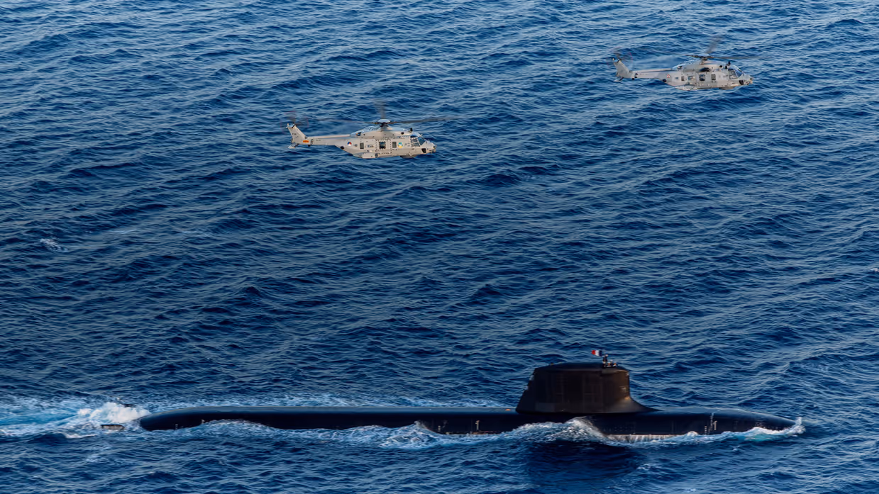 Een Franse onderzeeboot op volle zee, met daarboven 2 NH90’s.