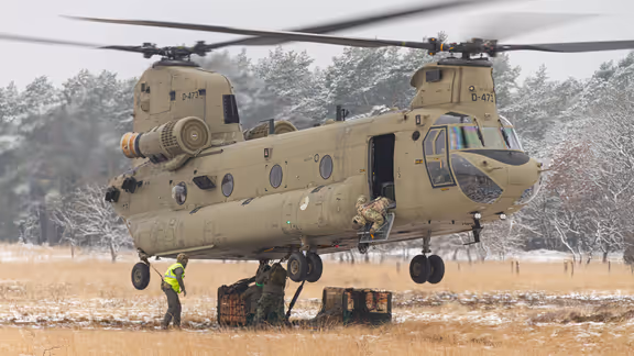 Een helikopter maakt een landing op een wit veld. Rechts met een paar pakketten onder zich. 