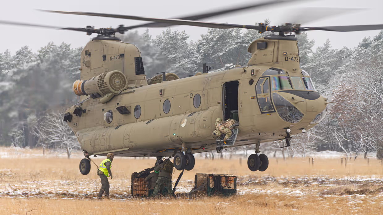 Een helikopter maakt een landing op een wit veld. Rechts met een paar pakketten onder zich. 