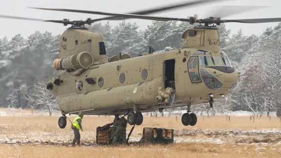 Een helikopter maakt een landing op een wit veld. Rechts met een paar pakketten onder zich. 