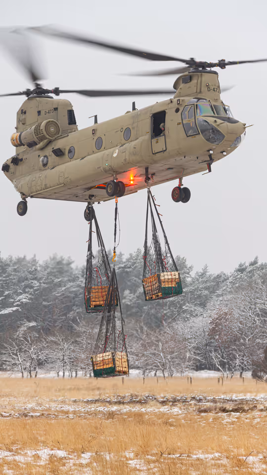 Een helikopter maakt een landing op een wit veld. Rechts met een paar pakketten onder zich. 