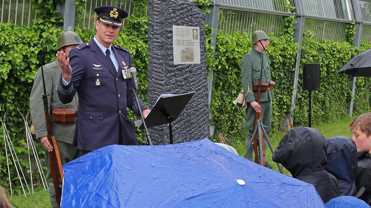 Een luchtmachtmilitair spreekt in de regen de aanwezigen toe. Op de voorgrond een grote blauwe paraplu met regendruppels erop. In de achtergrond het monument voor de omgekomen bemanningsleden van de vliegtuigen.