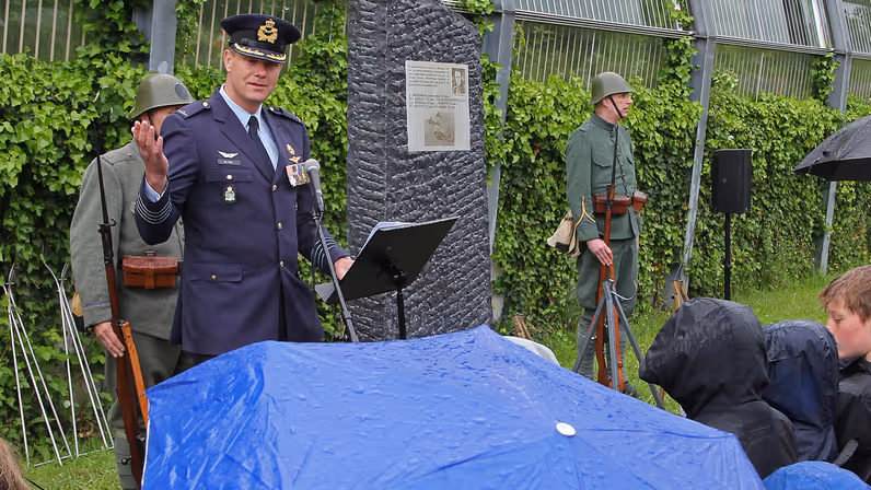 Een luchtmachtmilitair spreekt in de regen de aanwezigen toe. Op de voorgrond een grote blauwe paraplu met regendruppels erop. In de achtergrond het monument voor de omgekomen bemanningsleden van de vliegtuigen.