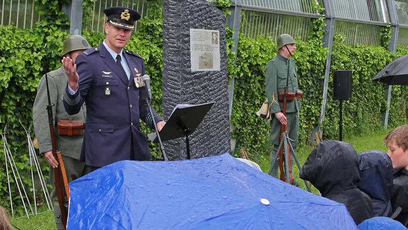 Een luchtmachtmilitair spreekt in de regen de aanwezigen toe. Op de voorgrond een grote blauwe paraplu met regendruppels erop. In de achtergrond het monument voor de omgekomen bemanningsleden van de vliegtuigen.