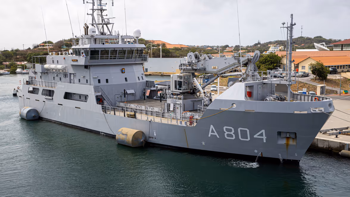 Zr.Ms. Pelikaan in een haven, omringd door water, gebouwen en heuvels.