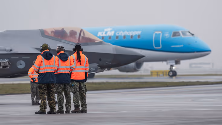 Militair personeel in oranje jassen bij een F-35 op een platform, met een KLM-vliegtuig op de achtergrond.